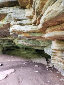 Layered rock formations showing sedimentary patterns with an overhanging ledge and a shallow cave-like opening at the base, surrounded by dry soil and scattered stones.