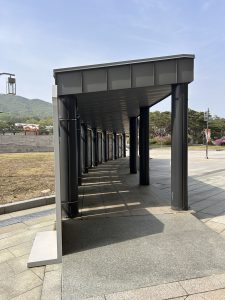 A long, covered walkway supported by dark grey columns. The overhead structure is metallic and slightly curved, and it casts shadows onto the light-colored, textured pavement below. In the background, there are rolling green hills and a clear blue sky, with a few trees and a light post visible on the right.