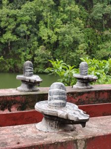 Three stone sculptures resembling Shiva Lingams are arranged on a weathered red platform, with lush green trees and a body of water visible in the background