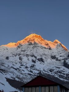 A snow-capped mountain peak illuminated by the golden light of sunrise, rising sharply against a clear blue sky. 