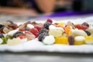 A close-up image featuring an assortment of colorful gummy candies scattered across a white surface. The candies come in various shapes and sizes, including bear-shaped and fruit-shaped gummies, with bright colors