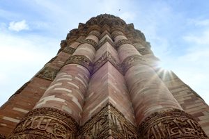 Qutb Minar, Delhi — a UNESCO World Heritage Site located in the Mehrauli area of South Delhi, India. It is a towering structure made of red sandstone, featuring intricately carved details along its base. The perspective is from the ground looking upward, showcasing the height of the pillar and its decorative elements. The sky above is partly cloudy, with a slight sunflare visible near the top of the pillar.