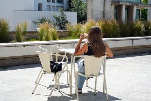 A person with long hair is sitting at a table outdoors, facing away from the camera and sipping a drink. They are wearing a black tank top and light blue jeans. A black bag is resting on a nearby chair, and there are green plants in the background, along with modern buildings.