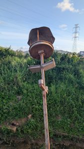 A rusty metal street light stands upright in a grassy area, featuring a circular, flat top with a wider, cylindrical section above it.