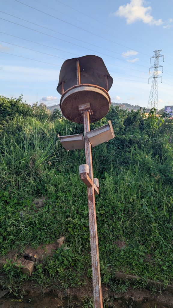 A rusty metal street light stands upright in a grassy area, featuring a circular, flat top with a wider, cylindrical section above it.