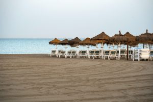 A serene beach scene featuring a row of white lounge chairs arranged under straw umbrellas. The sandy beach stretches out in the foreground, while the calm, blue ocean meets a clear sky in the background.