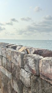 A close-up view of a stone wall made of large, rough-hewn rocks, with a gap in the middle. Beyond the wall, a calm ocean stretches to the horizon under a partly cloudy sky, with a few small boats visible in the distance. The scene has a serene, coastal atmosphere.
