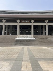 A large building with a prominent staircase leading up to it. The structure features traditional architectural elements, including decorative columns. Above the entrance, there is a sign with Korean text. The area in front of the building is paved with patterned stones, and a few people can be seen near the entrance.