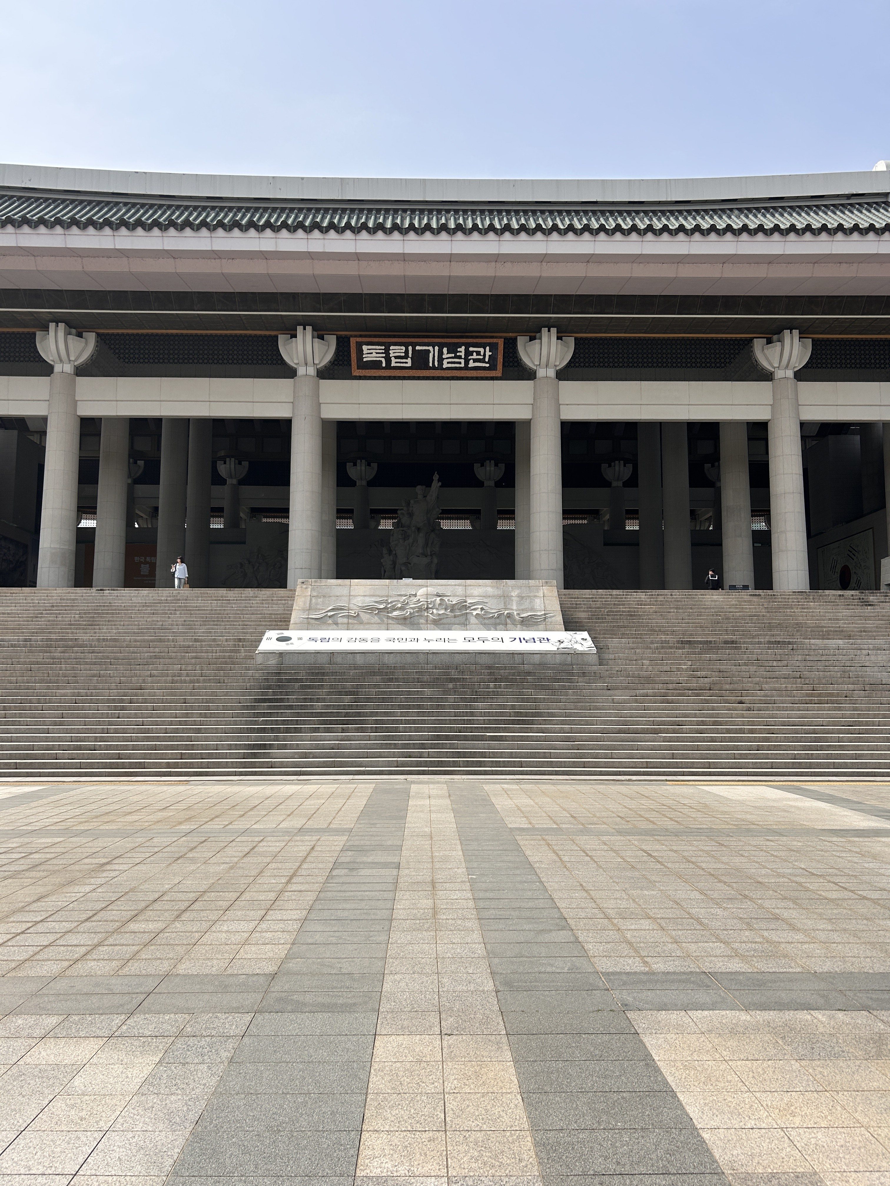 A large building with a prominent staircase leading up to it. The structure features traditional architectural elements, including decorative columns. Above the entrance, there is a sign with Korean text. The area in front of the building is paved with patterned stones, and a few people can be seen near the entrance.
