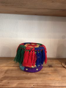 A round, colorful woven container adorned with vibrant patterns and fringes of red, green, and blue. 
