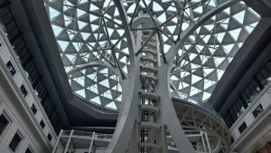 The image showcases a modern architectural interior featuring a geometric glass ceiling with a triangular pattern. Below the ceiling, a spiral structure made of white material rises vertically, flanked by surrounding walls that include large windows.