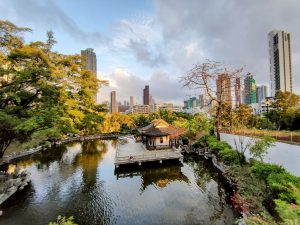 

A peaceful traditional Chinese pavilion park scene featuring a serene pond surrounded by lush greenery and trees. In the center, there is a traditional pagoda-style structure with an ornate roof, situated on a small island. Behind the pond, modern skyscrapers rise against a partly cloudy sky, creating a contrast between nature and urban architecture.