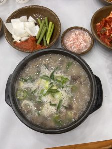 A black stone bowl containing a steaming soup with floating pieces of meat and garnished with chopped green onions. Surrounding the bowl are several small brown dishes, one with sliced white onions, green onions, and a dollop of red chili paste, another with a pink seafood item, and a dish of kimchi with red pepper seasoning. 