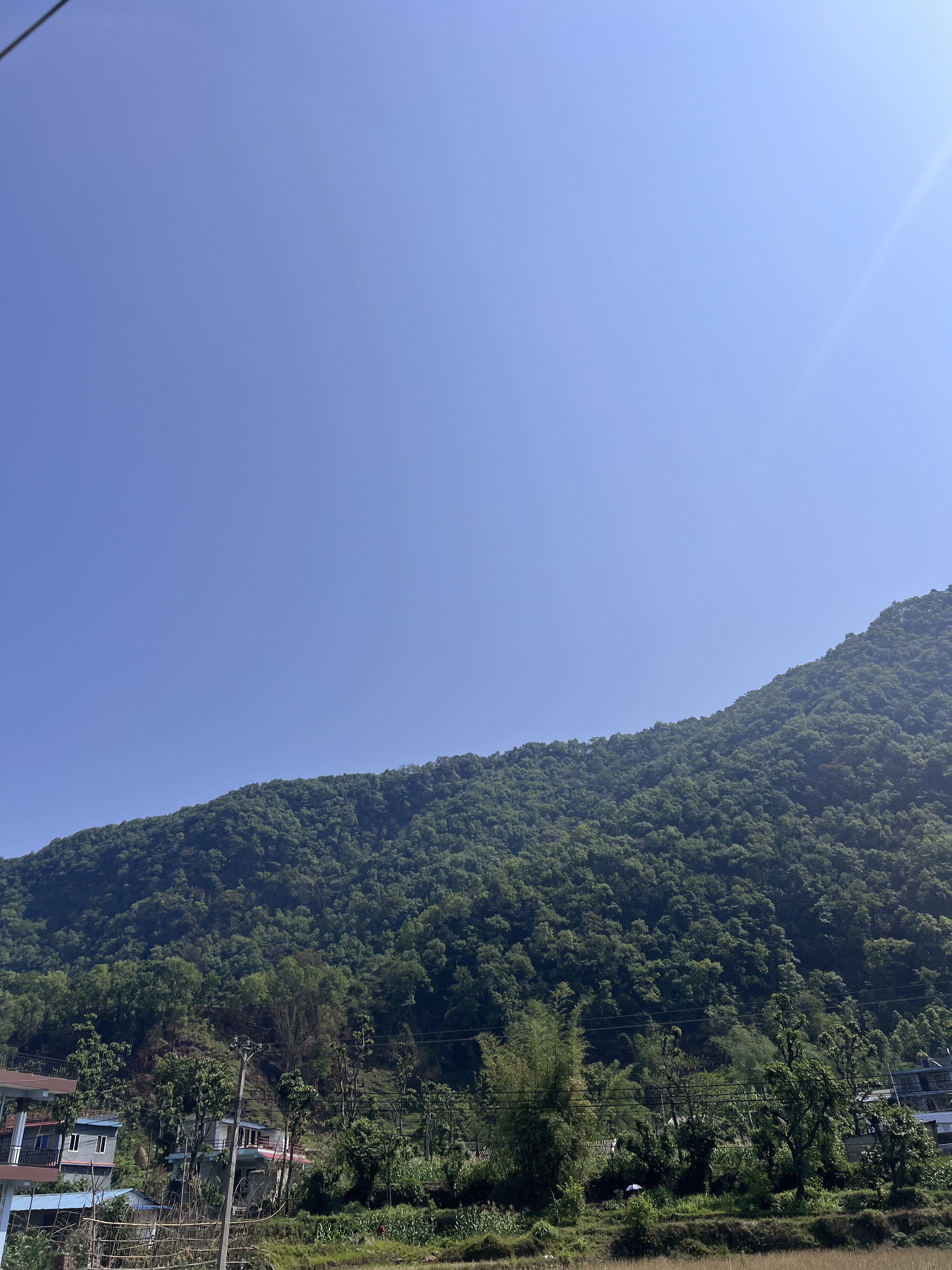 A clear blue sky is visible above a lush green mountain landscape. The lower portion of the image features a few buildings, likely residential, surrounded by trees and vegetation.