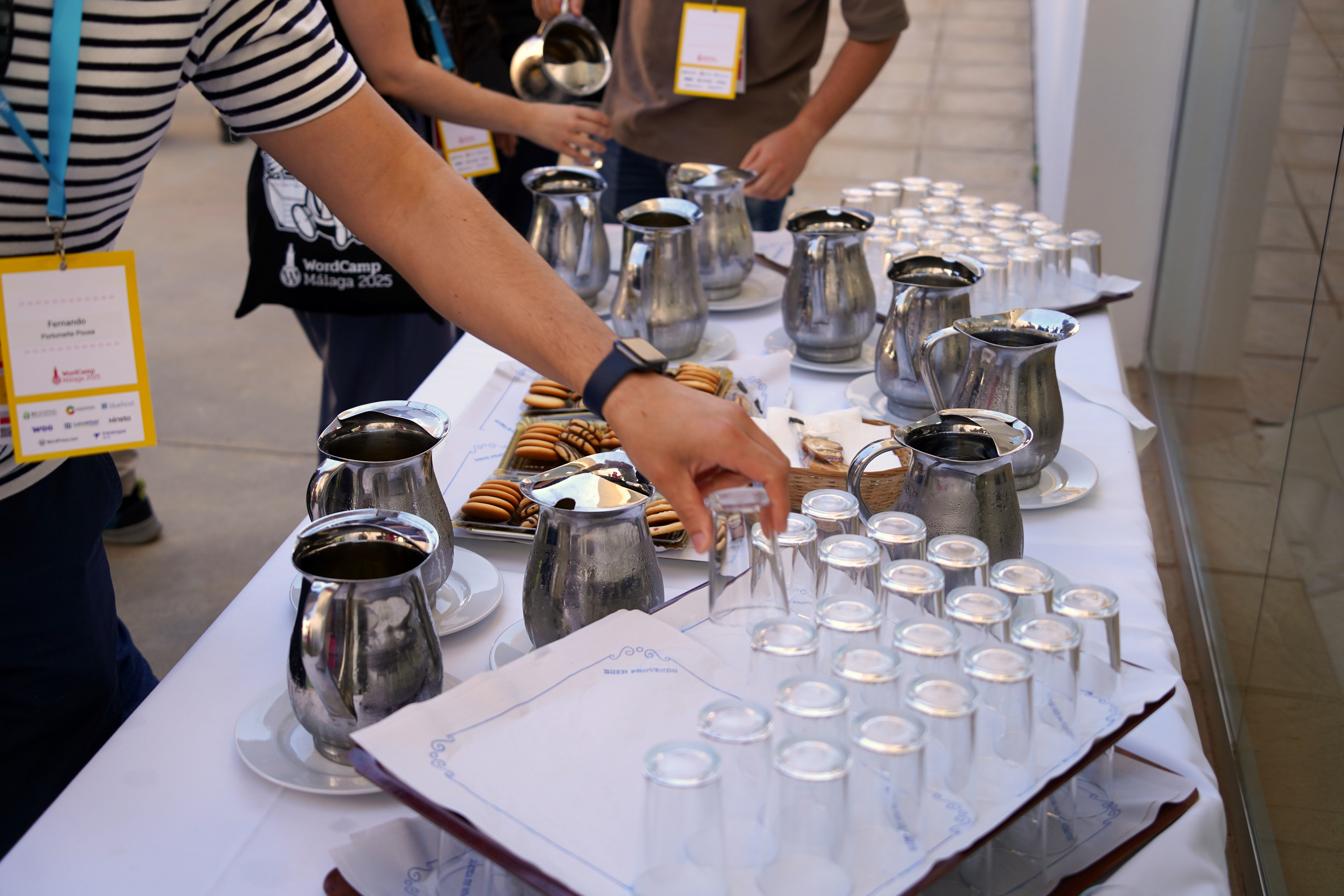 A table is set up with multiple silver pitchers and several transparent glasses arranged neatly in rows. In the foreground, a person is reaching for a glass, while another individual in the background is holding a pitcher. There are also plates of cookies on the table.