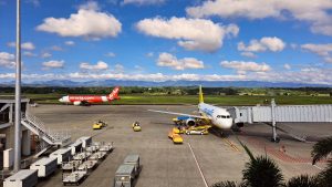 Commercial plane taxiing and parked for passengers embarking with a mountain range as background under sunny blue sky.
