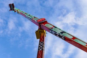 A tall amusement ride in an amusement park stretches upwards against a bright blue sky with scattered clouds. The ride features a colorful design with red, green, and checkered patterns, and a round sign at the top that reads "GIGANT".