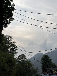 View of forested hilltops in the distance, against a cloudy sky. Many power cables crisscross the sky.
