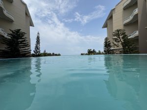 A serene view of an infinity pool reflecting the sky, flanked by tall, slender pine trees and the side of a beige building. In the distance, the ocean is visible under a partly cloudy sky.
