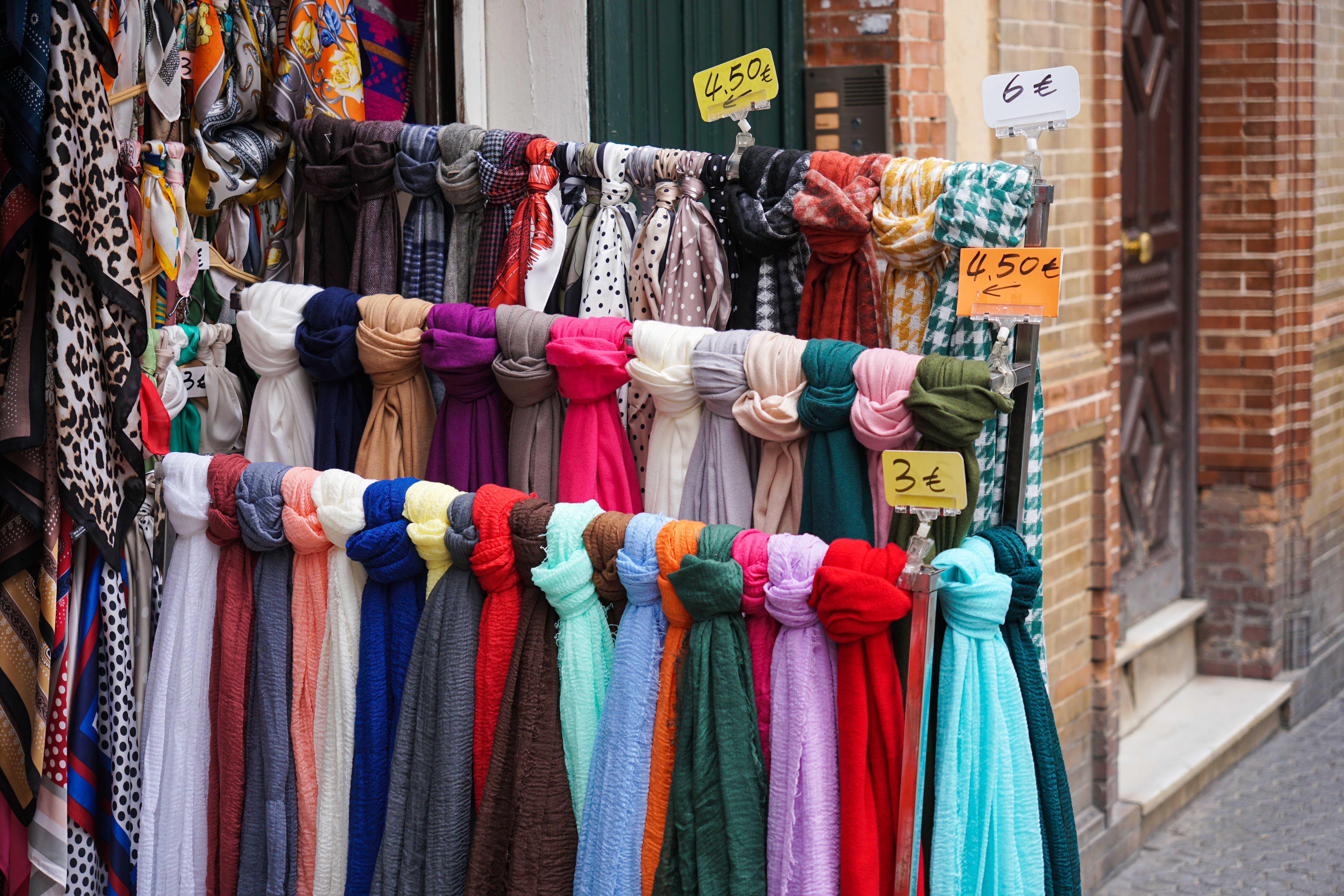 A display of colorful scarves for sale, hung on a rack outdoors. The scarves are arranged in vibrant hues, including shades of purple, pink, red, blue, green, and white. Some of the scarves are patterned, while others are solid colors. Various price tags are visible