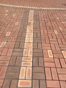A close-up view of a patterned brick pavement, featuring alternating colors of reddish-brown and light tan bricks. Some areas show signs of wear, with grass growing between the crack
