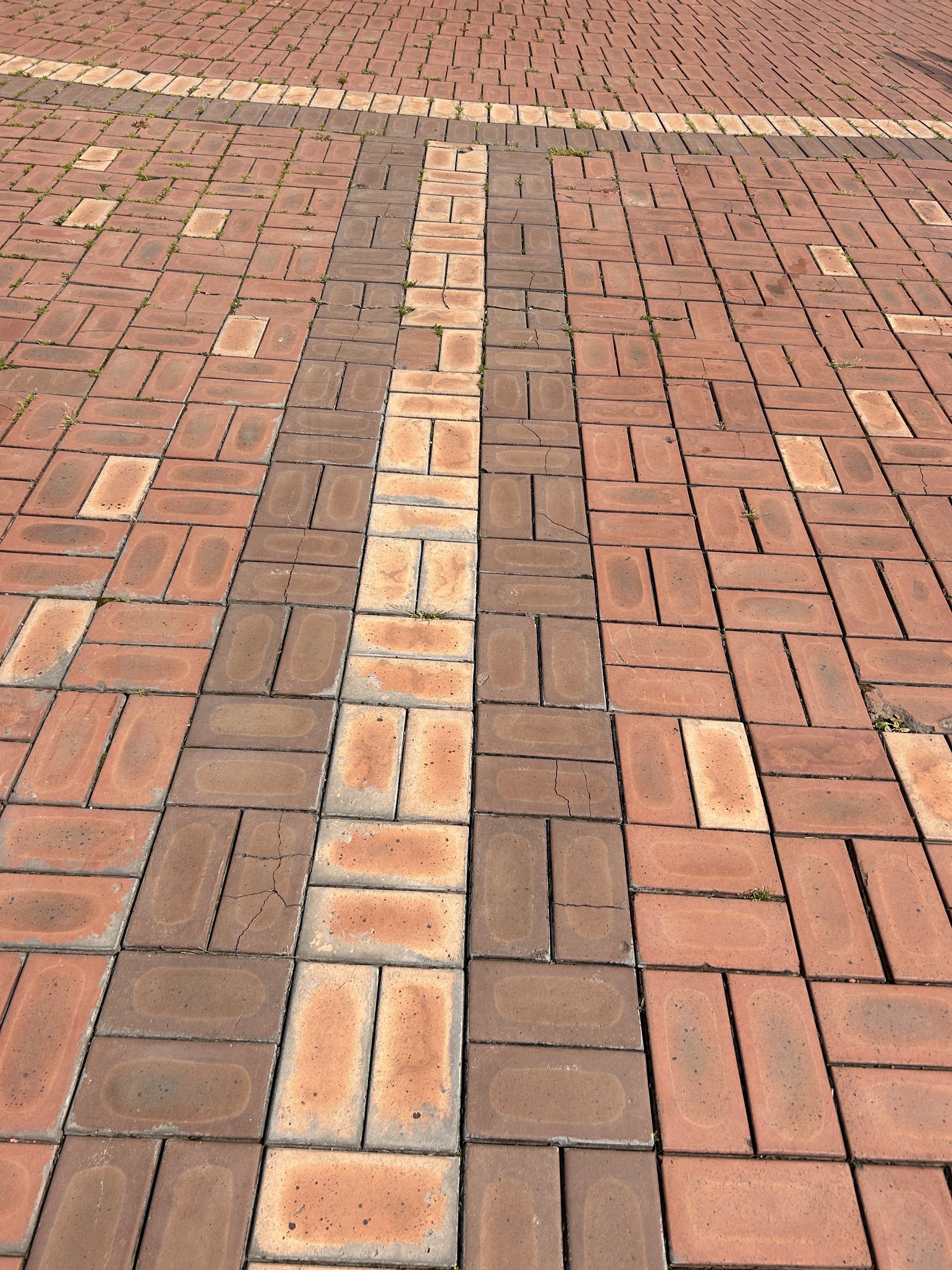 A close-up view of a patterned brick pavement, featuring alternating colors of reddish-brown and light tan bricks. Some areas show signs of wear, with grass growing between the crack