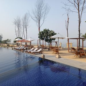 A serene poolside scene featuring an infinity pool with dark blue tiles, surrounded by wooden lounge chairs and white umbrellas. In the background, there are bare trees and lush greenery, along with wooden structures resembling swings and shaded sitting areas.
