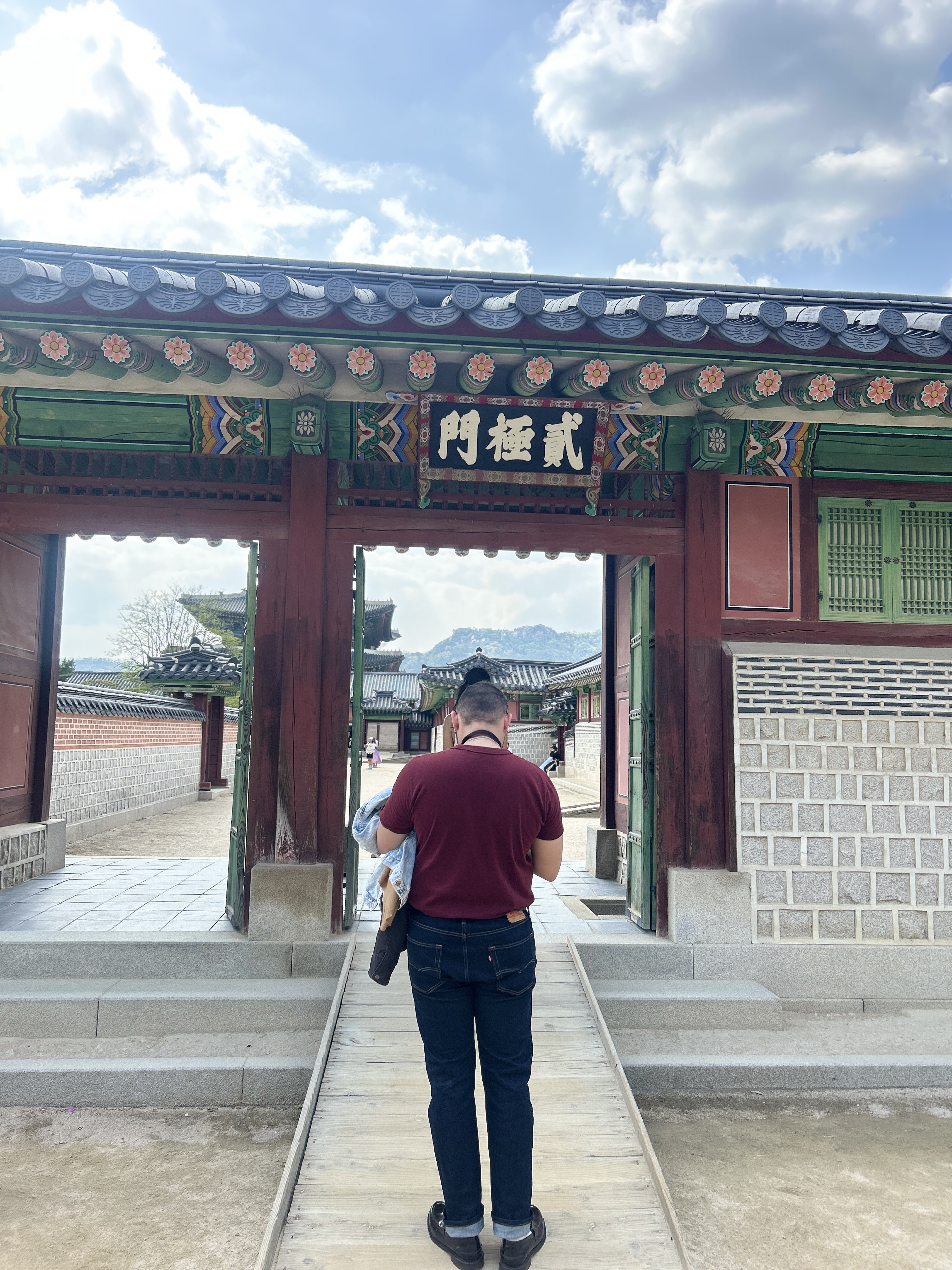A person in a maroon shirt stands facing a traditional wooden gate adorned with colorful decorations and Chinese characters