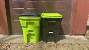 A pair of large plastic rubbis bins in front of a garage door. On is green with a black lid, and the other is black with a green lid, one for trash and one for recycling.