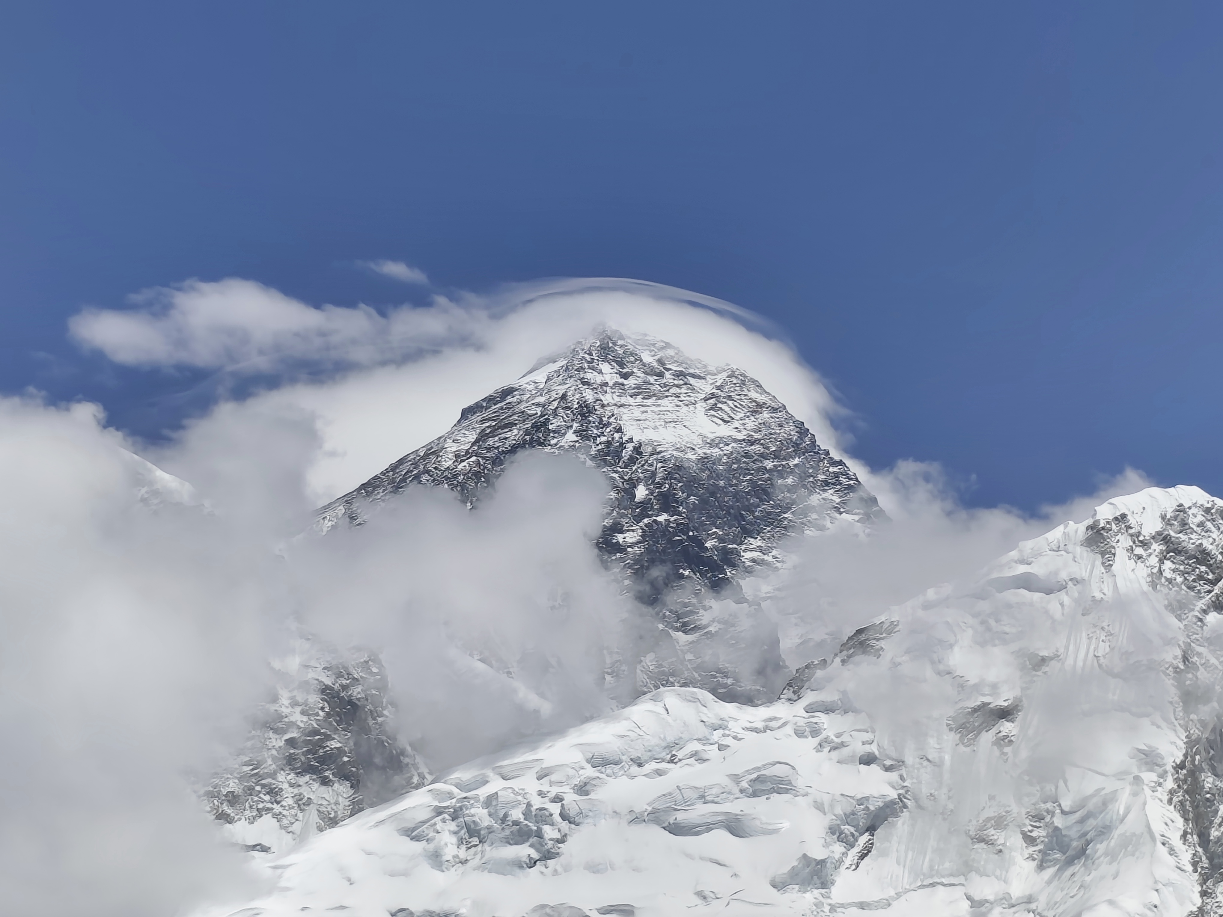 A majestic snow-capped everest mountain peak rises into a clear blue sky, surrounded by wispy clouds.