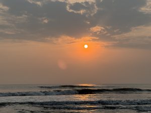 A serene ocean view at sunset, with the sun appearing as a bright orange orb partially hidden behind soft clouds
