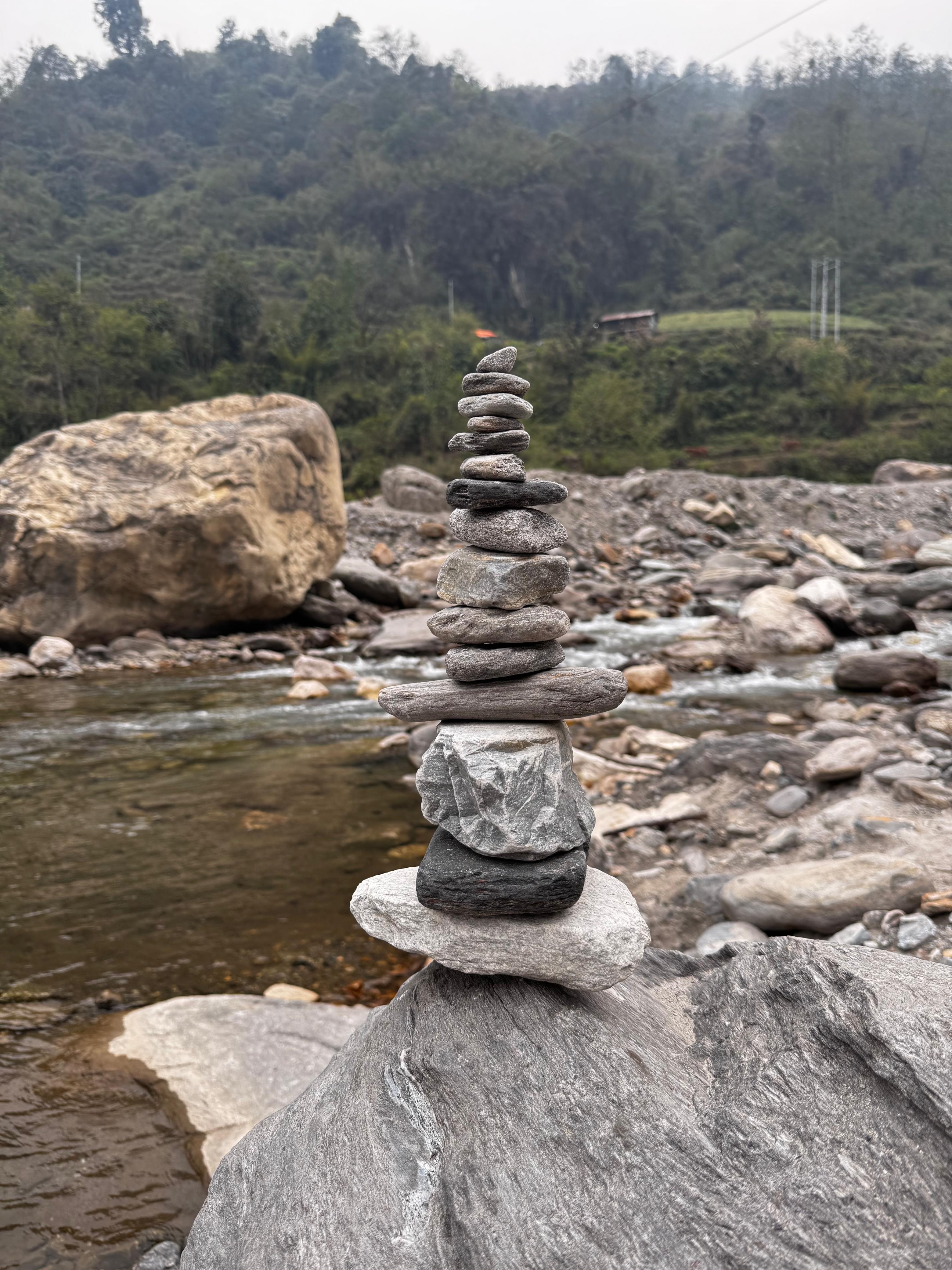 A stack of smooth, rounded stones balances precariously in the foreground, set against a rocky riverbed with a shallow stream flowing nearby.
