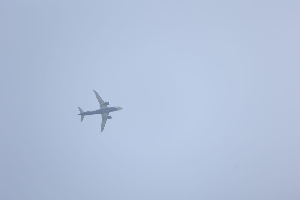 An airplane flying in a clear blue sky, viewed from below, with its wings extended and a light gray background.