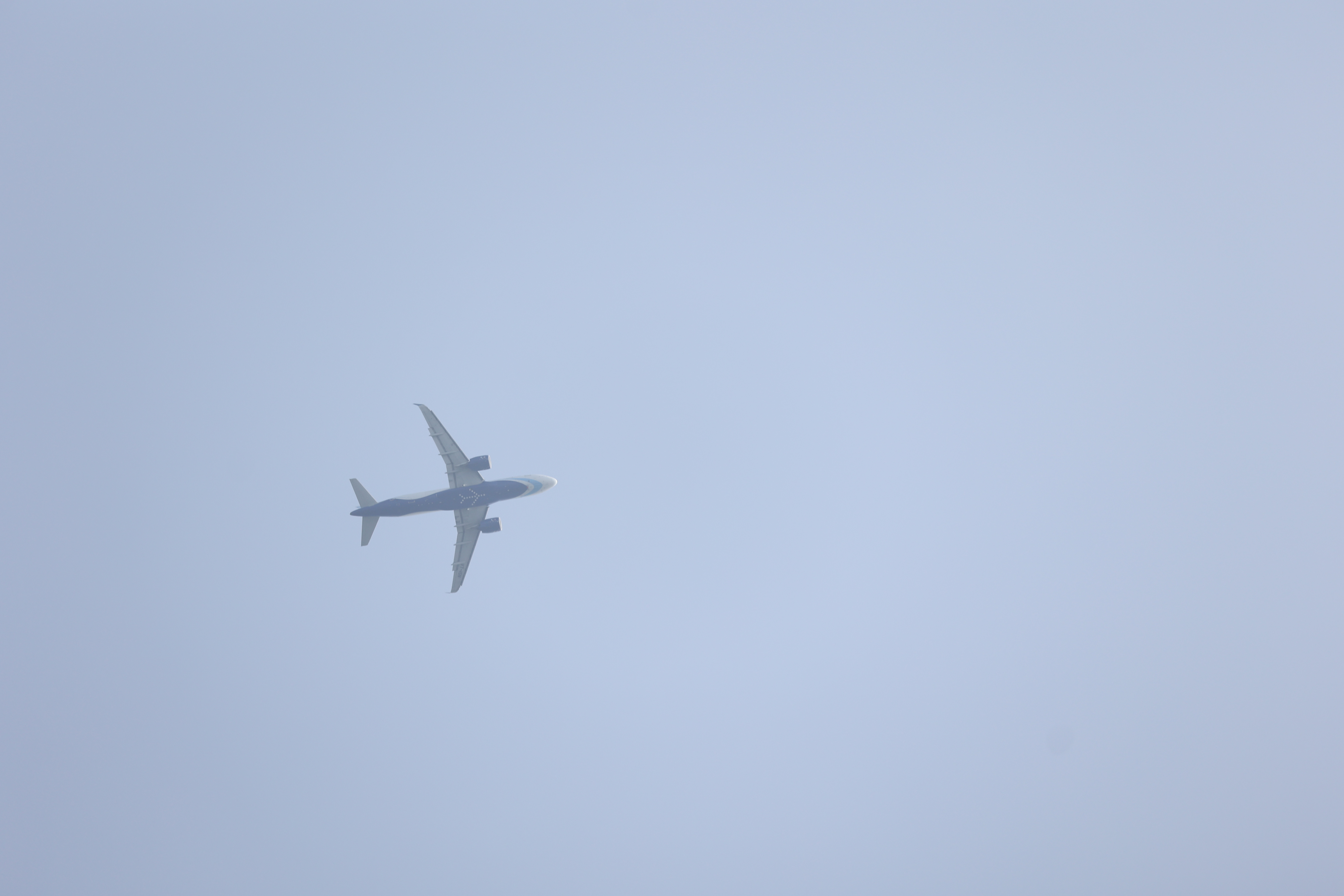 An airplane flying in a clear blue sky, viewed from below, with its wings extended and a light gray background.