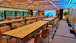 A spacious conference room featuring multiple rows of rectangular wooden tables surrounded by beige chairs