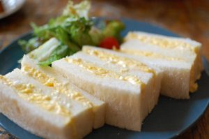 Egg sandwich served on soft white bread, filled with creamy yellow egg mixture, paired with a fresh side salad of leafy greens and a cherry tomato
