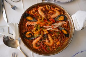 A large paella pan filled with a colorful seafood dish featuring shrimp, mussels, and an array of vegetables such as bell peppers and chives, resting on a white tablecloth. A silver serving spoon and fork are partially visible alongside a folded napkin.