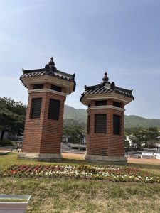 Two traditional-style brick structures with pointed roofs are positioned side by side in a green park. 