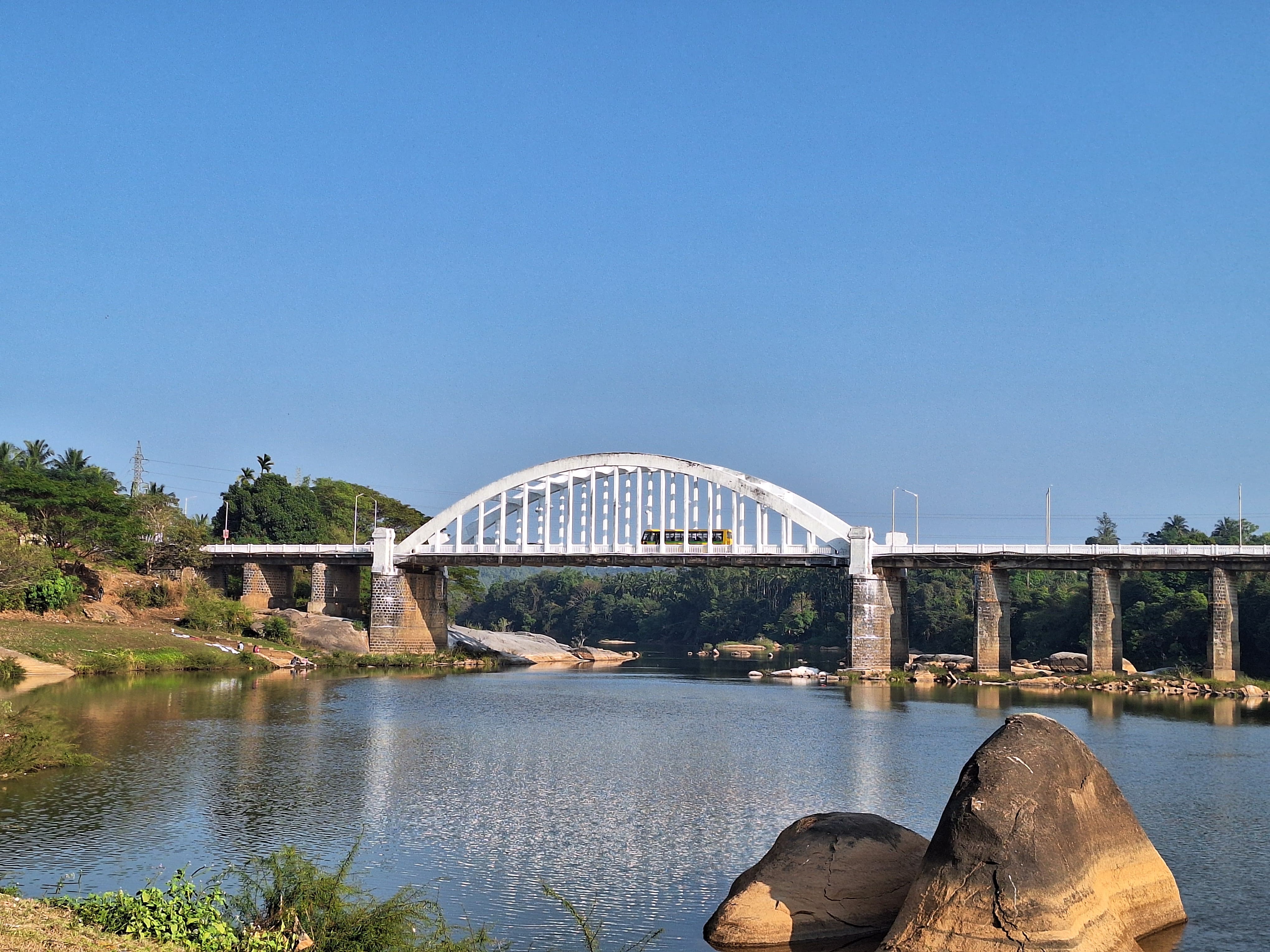 The image shows the Tunga Bridge, an old white bridge with an arched design, crossing the Tunga River in Thirthahalli, Karnataka. The bridge, designed by Sir M. Visvesvaraya, is framed by lush greenery on the riverbanks and large rocks in the foreground, all under a clear blue sky.