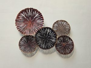 A collection of five decorative plates arranged on a light-colored wall. The plates vary in size and feature intricate cut-out designs with radiating patterns. The colors range from deep reds to dark browns and muted metallic shades, creating a visually striking contrast against the wall.