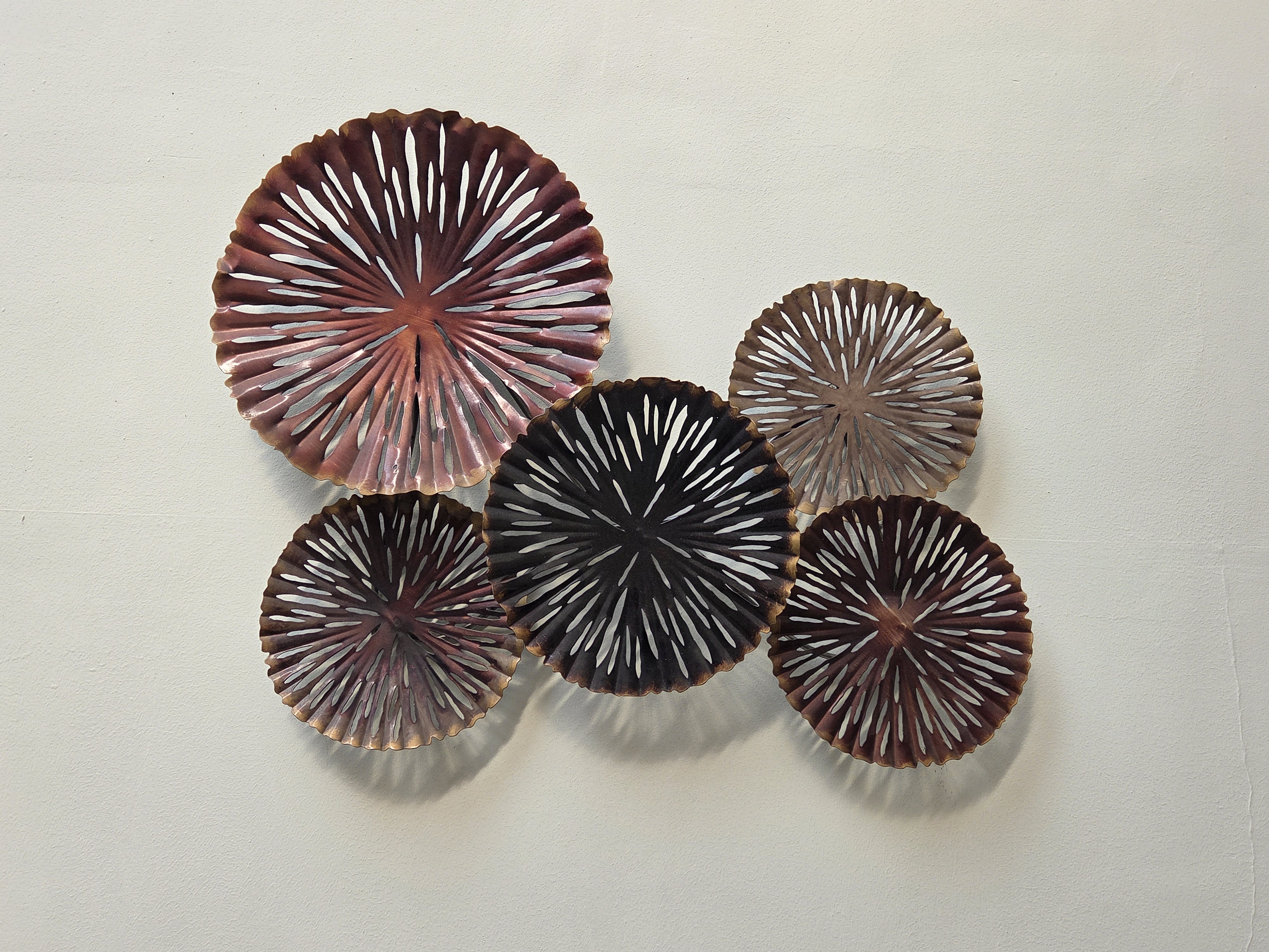 A collection of five decorative plates arranged on a light-colored wall. The plates vary in size and feature intricate cut-out designs with radiating patterns. The colors range from deep reds to dark browns and muted metallic shades, creating a visually striking contrast against the wall.