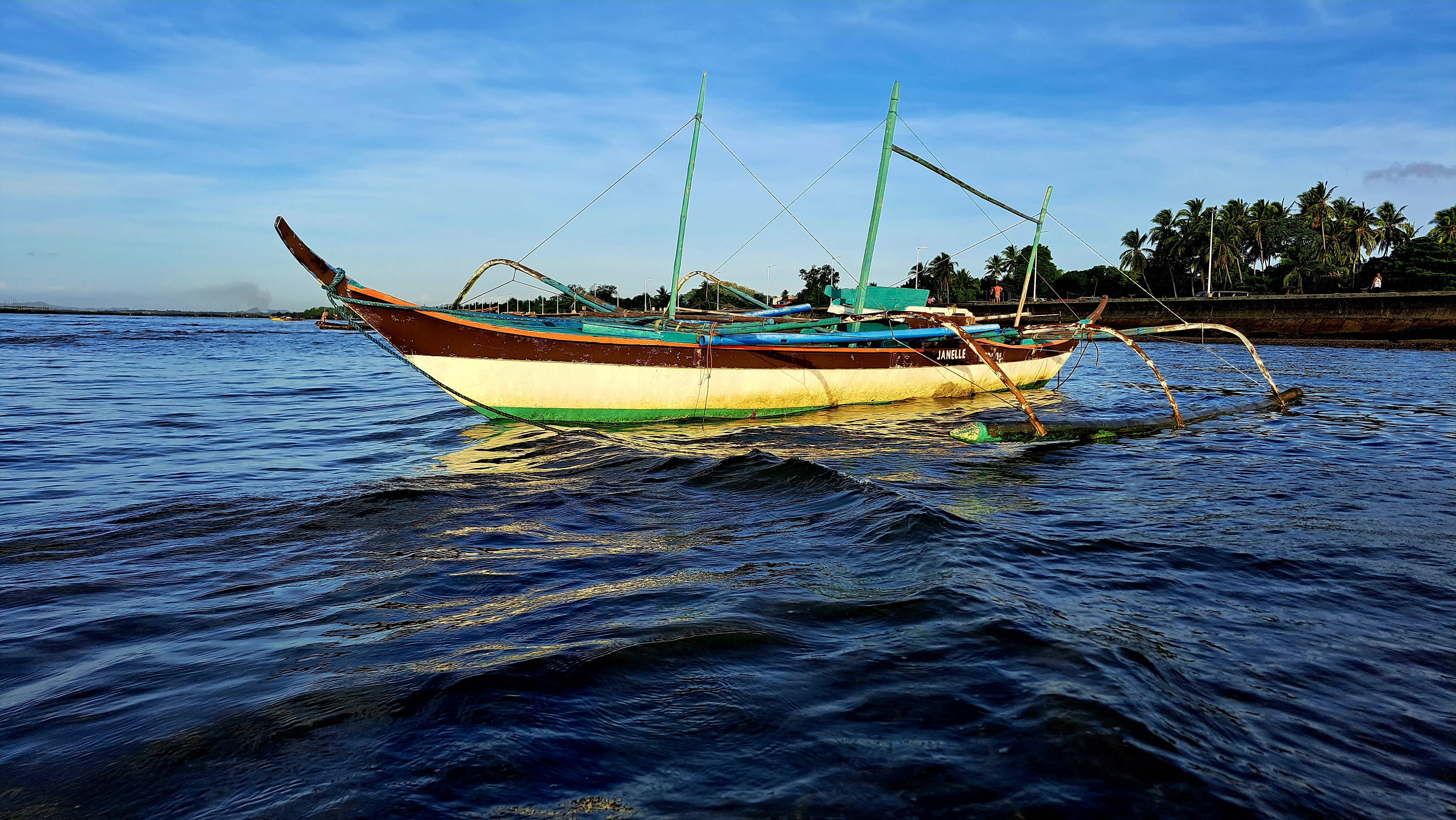 A wooden boat with a brown and white hull and green accents is anchored in calm waters. The boat features two outriggers and is tied with ropes.