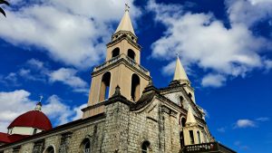 A historic church building with two prominent towers, the taller one featuring a pointed roof and a cross at the top.