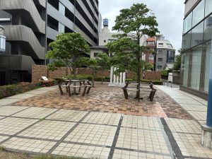 A landscaped outdoor area featuring a paved patio with square tiles, surrounded by low shrubs and small trees. In the center, there are several wooden benches arranged in a circular pattern under the shade of trees. 