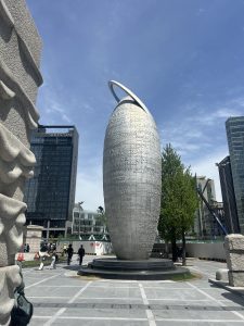A tall, abstract sculpture resembling an elongated oval with a circular ring on top is centrally positioned in an urban setting.
