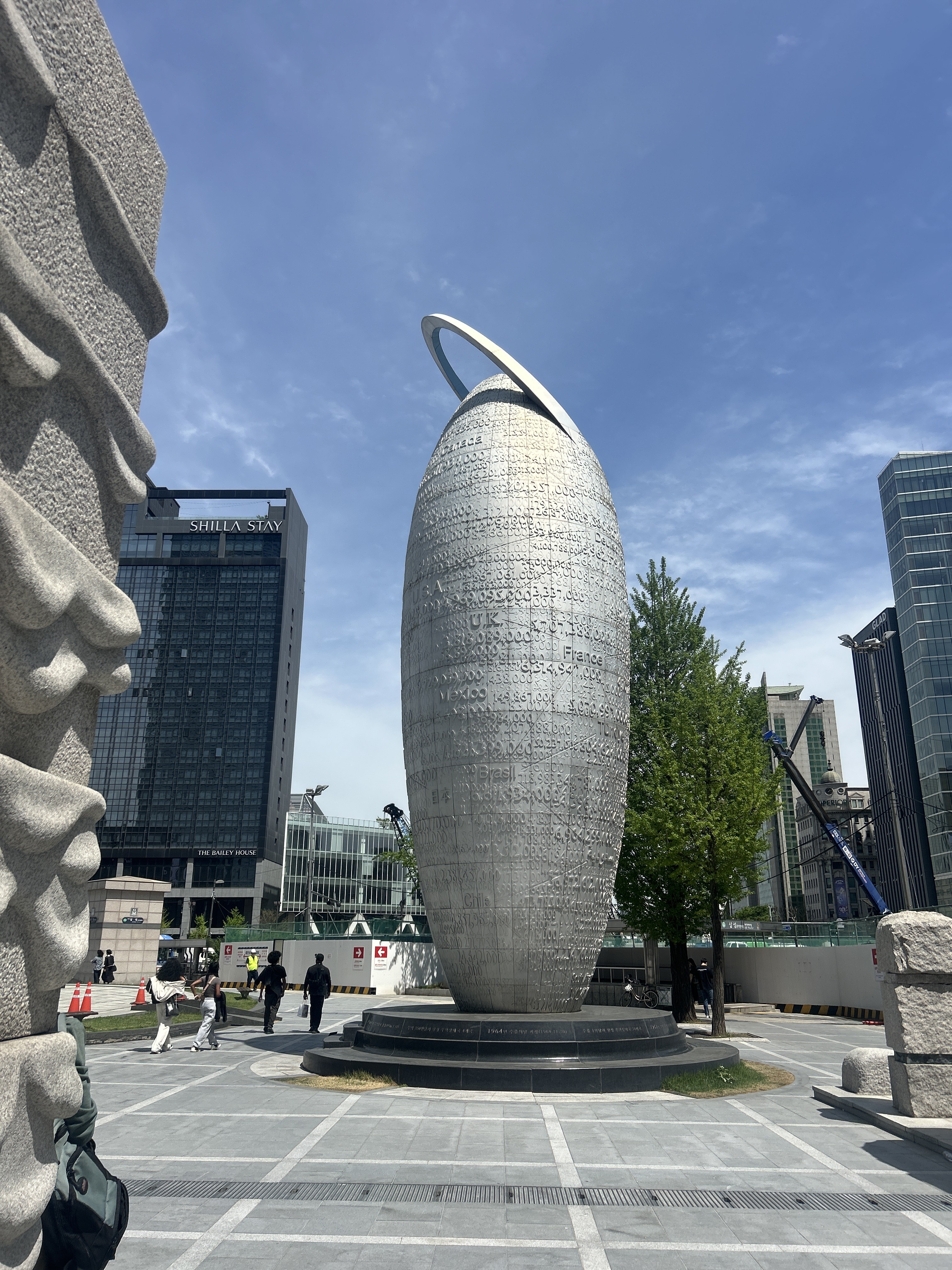 A tall, abstract sculpture resembling an elongated oval with a circular ring on top is centrally positioned in an urban setting.
