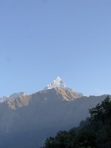 A majestic snow-capped mountain peak rises above a range of lower, rugged mountains, set against a clear blue sky