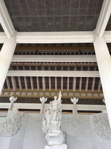 Architectural structure featuring a series of massive columns, a textured ceiling, and a prominent sculptural relief. In the foreground, a detailed statue group rises from a base, depicting figures in dramatic poses.