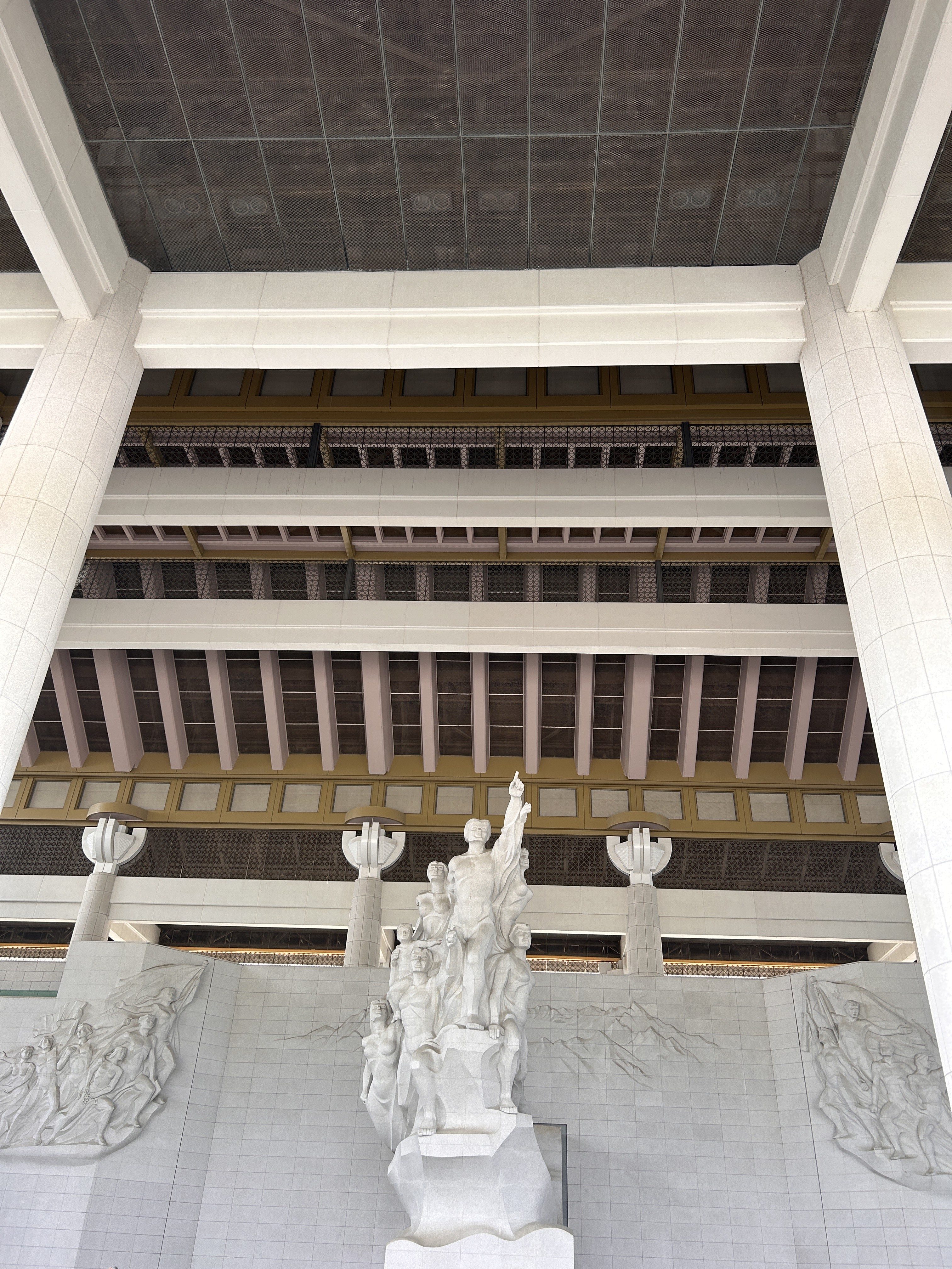 Architectural structure featuring a series of massive columns, a textured ceiling, and a prominent sculptural relief. In the foreground, a detailed statue group rises from a base, depicting figures in dramatic poses.