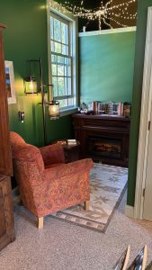 A nook in a book shop, about 1.5 meters square with a fireplace in the back, a large window on one wall, and a big comfy chair facing into the nook. There are tiny white lights on the ceiling. 